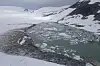 Aerial view of an ice-filled meltwater lake surrounded by snow and rocky slopes.
