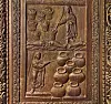 Brown carved panel relief featuring figures, bowls, and jars within a decorated frame.