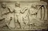 Stone relief featuring a central naked figure, people, animals, and columns.